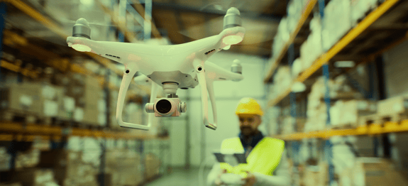 Image of warehouse worker standing in warehouse and operating drone, a banner image for Routeique's blog on drones in the ware